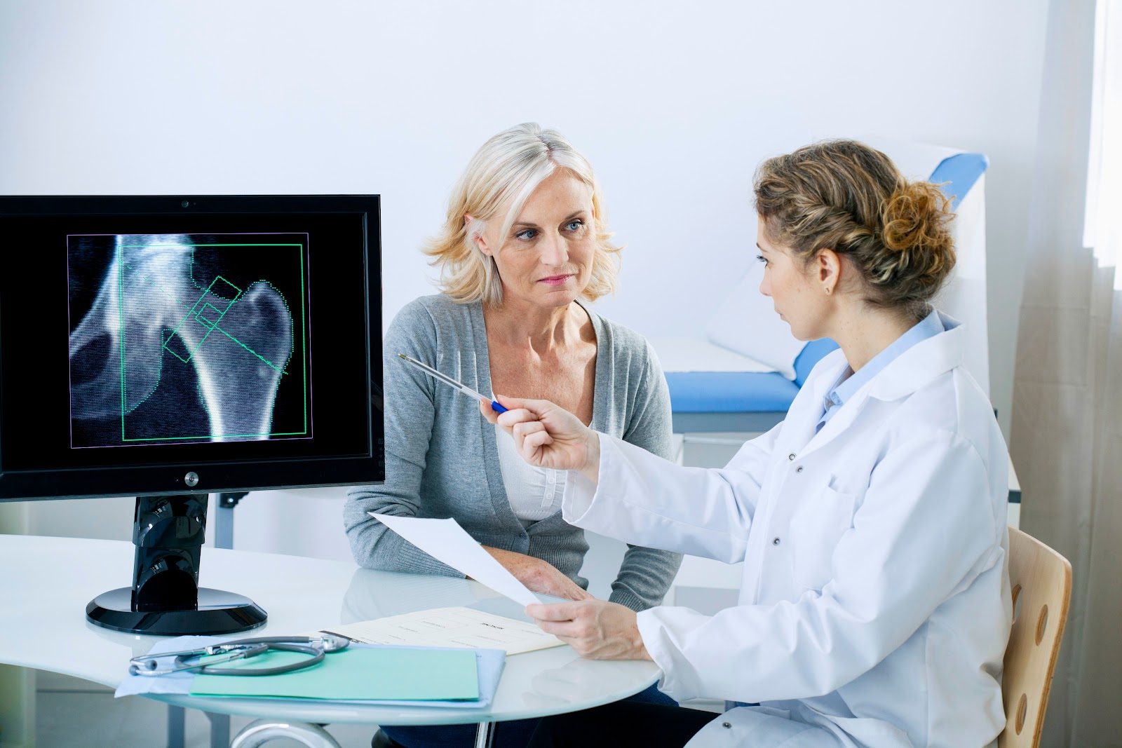Clinician in a white coat seated beside an older woman patient, pointing with a pen at a monitor displaying a hip bone-density (DEXA) scan with green measurement regions marked on the femoral neck