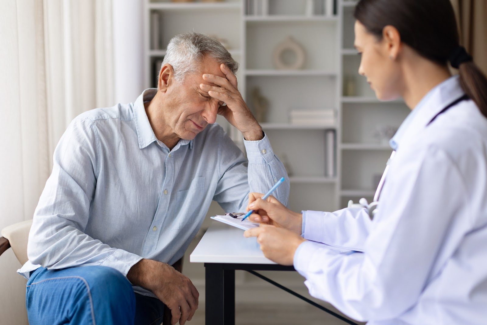 An older man in a light shirt resting his head in his hand across the desk from a female physician who takes notes on a clipboard — the fatigued patient that Nutrition 244 teaches chiropractors to evaluate and manage with chiropractic care and nutrition