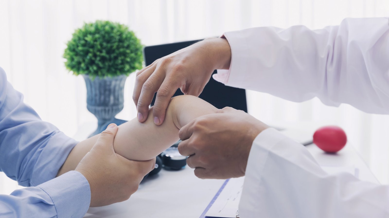 A clinician's hands palpate a seated patient's wrist while stabilizing the closed hand in a bright consultation room — the hands-on orthopedic testing taught in Physical Examination 214: Wrist, Hand and Thumb, a two-hour chiropractic CE course from CCEDseminars