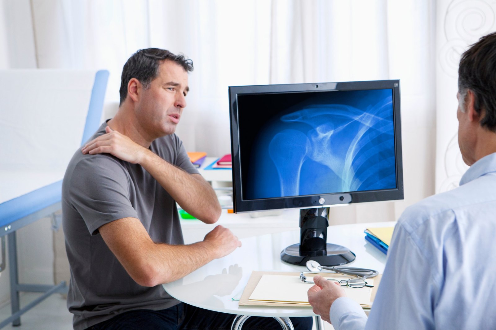 Patient in a gray T-shirt holding his painful shoulder and describing the complaint while a clinician in a white coat reviews a shoulder radiograph on a monitor between them — the shoulder complex examination, the subject of Physical Examination 209: Shoulder Complex, a two-hour chiropractic CE course from CCEDseminars