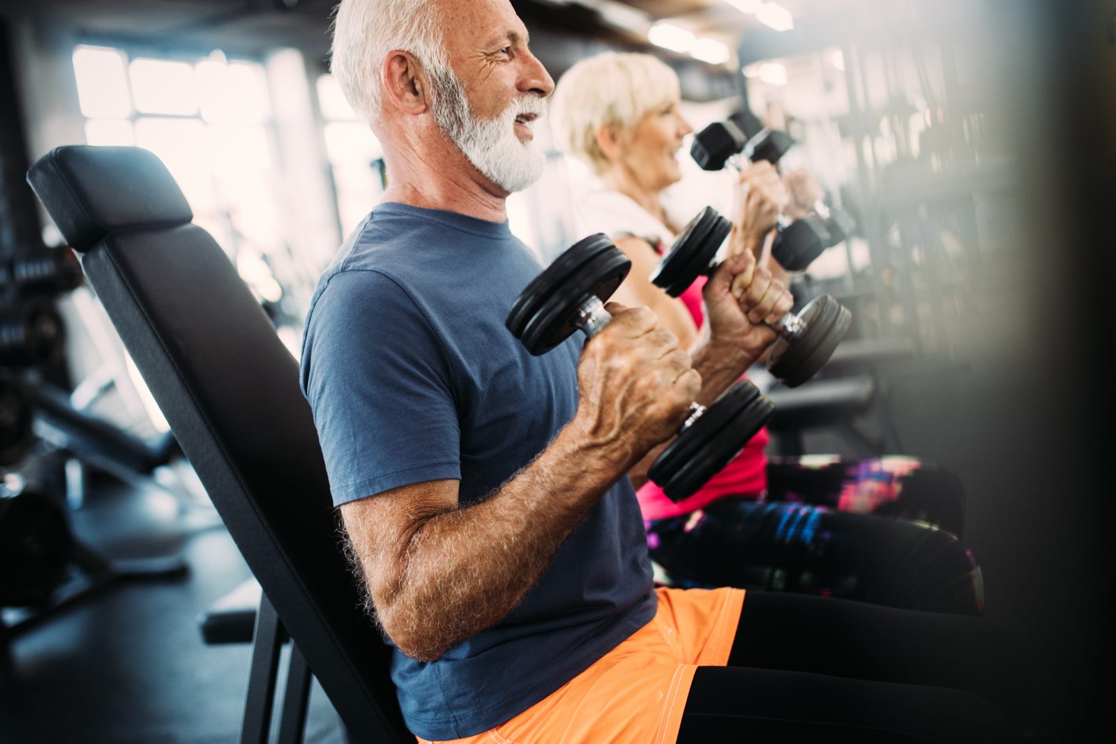 Older man and woman performing seated dumbbell curls side by side in a gym, illustrating supervised strength training for older adults