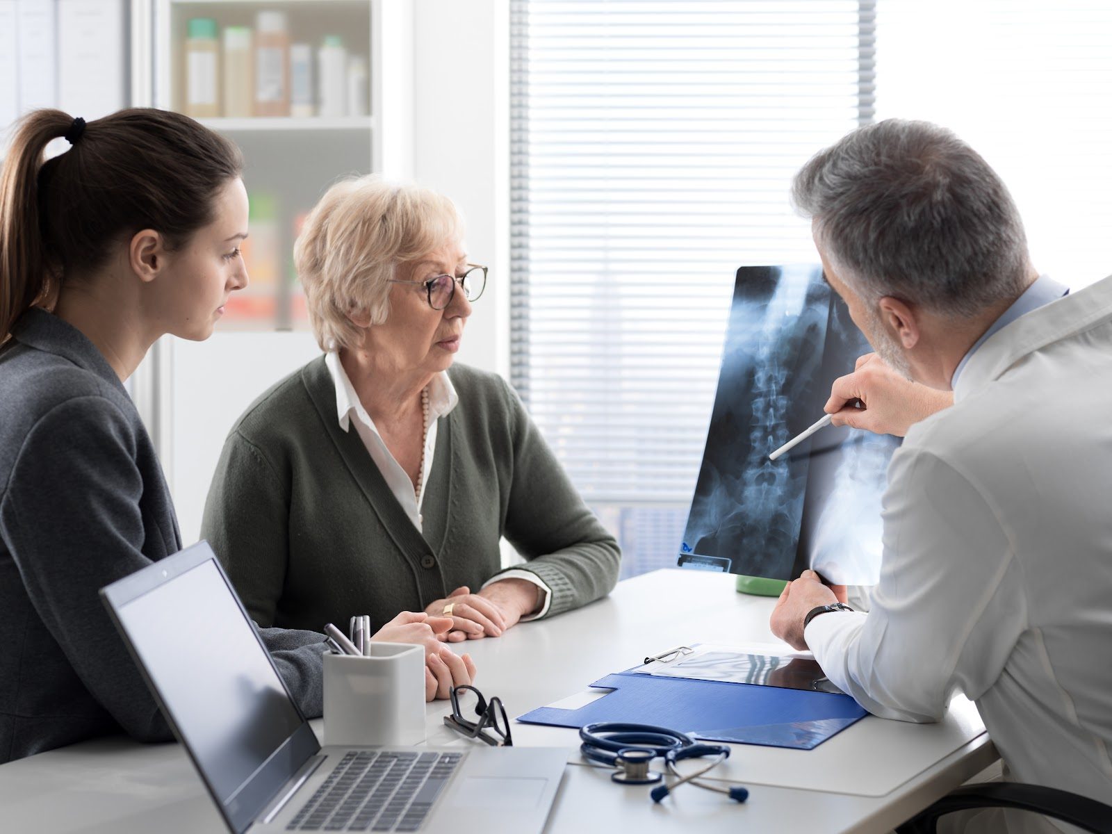 An older woman with glasses consulting with a clinician who holds up an X-ray while a younger woman looks on — the everyday sprains, strains, tendonitis and bursitis that Nutrition 239 teaches chiropractors to manage with supplements and anti-inflammatory nutrition