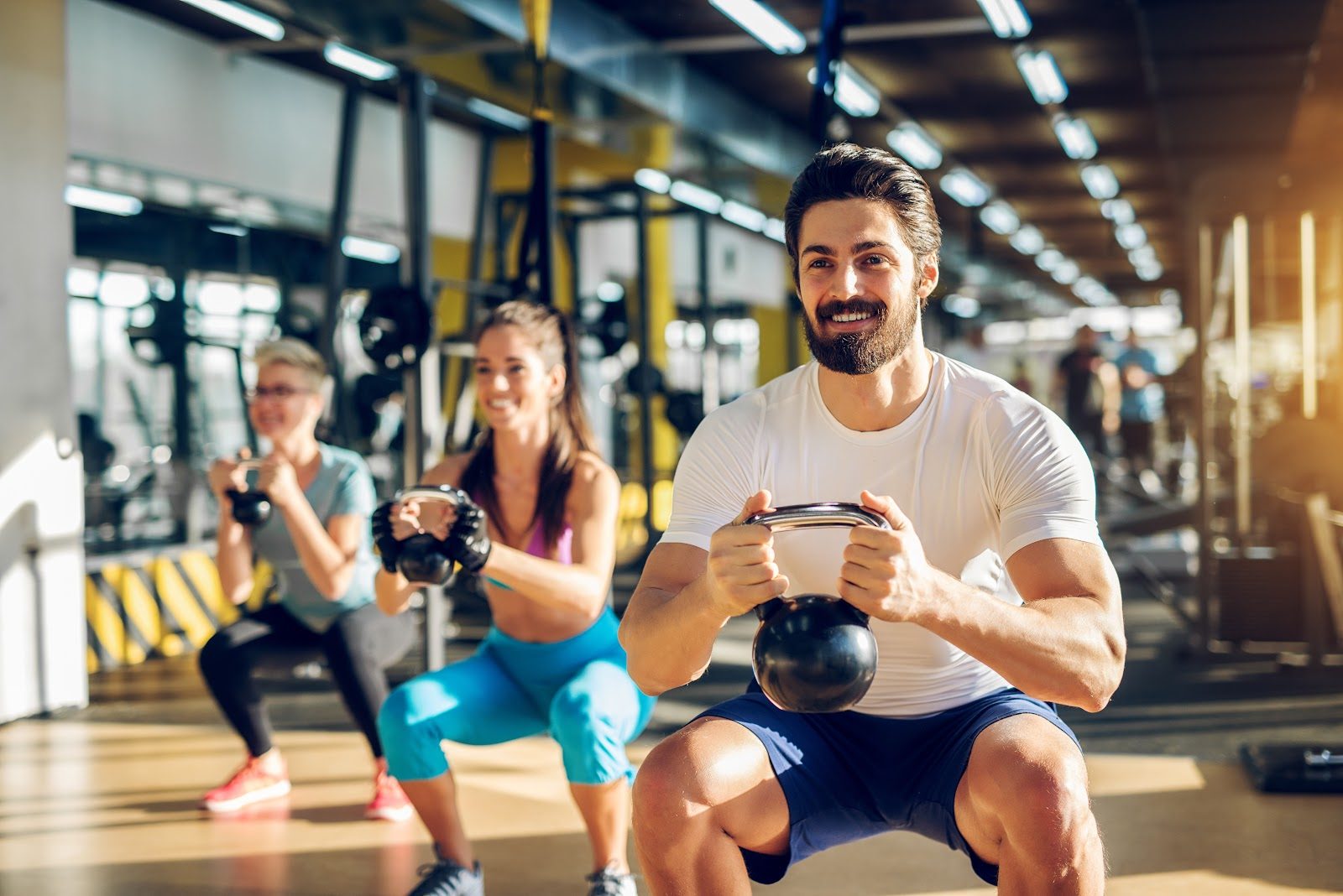 Group exercise session in a modern gym, a smiling bearded man in a white shirt and two women performing kettlebell squats on a wooden floor