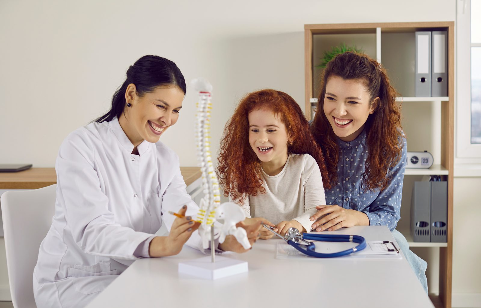 Smiling clinician showing a spine model to a young girl and her mother at a consultation table — Pediatrics 201: Introduction to Chiropractic Pediatrics, a 2-hour chiropractic CE course