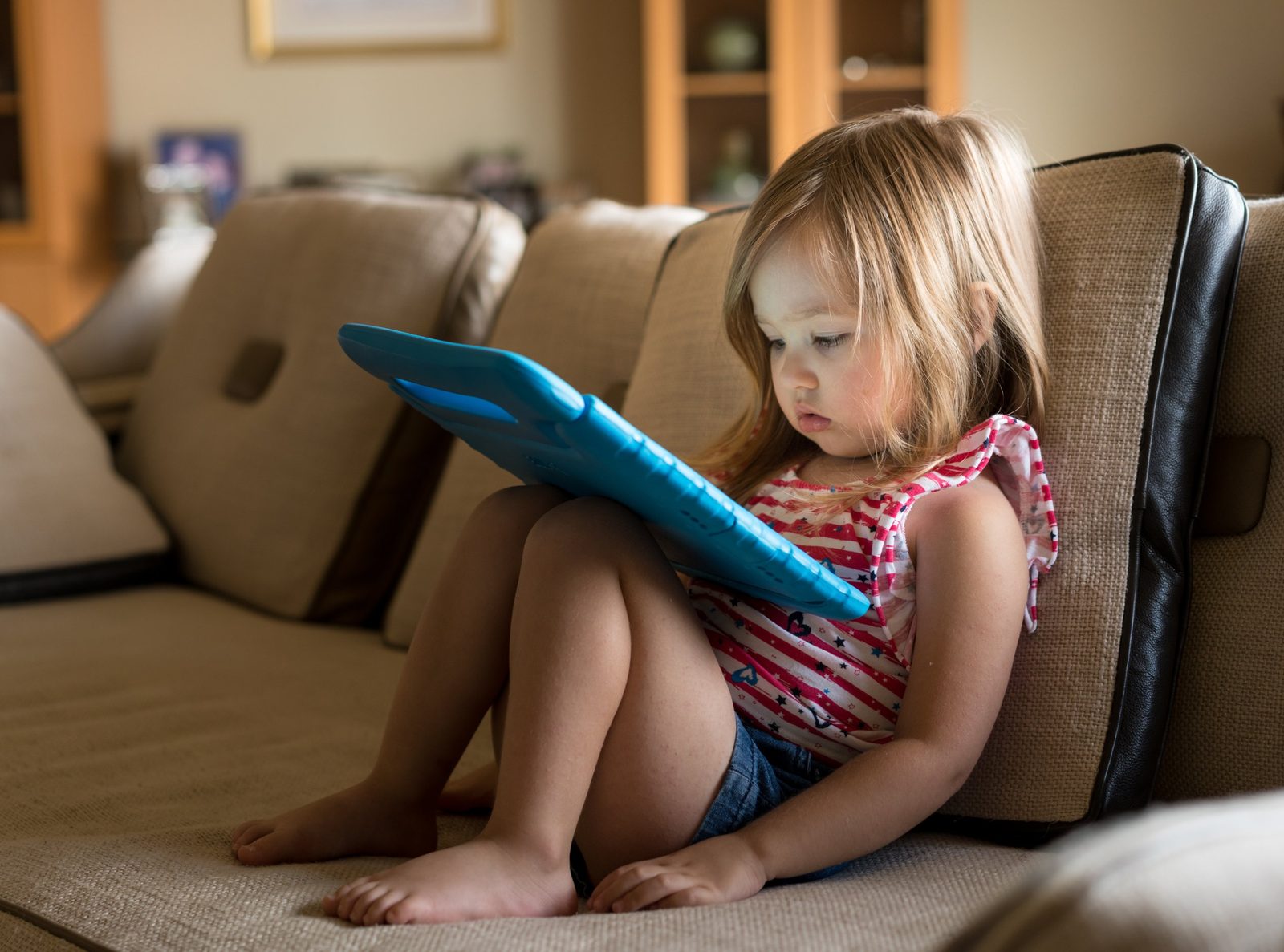 Toddler girl in a striped tank top sitting barefoot on a beige sofa, head bent forward over a tablet in a blue child case — the flexed-neck screen posture examined in Pediatrics 215, a pediatric text neck chiropractic CE course from CCEDseminars