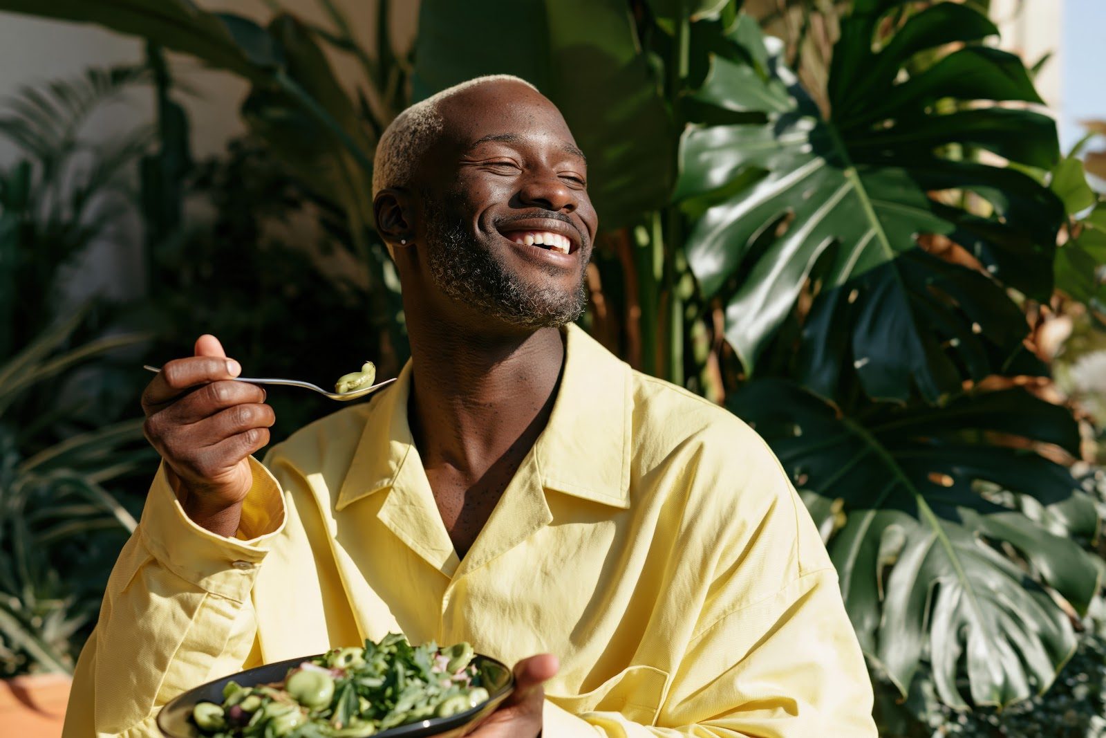 A smiling man in a yellow shirt eating a fresh salad among tropical greenery — the mood and stress resilience that Nutrition 219 teaches chiropractors to support with advanced neurotransmitter, HPA-axis and gut-brain nutrition