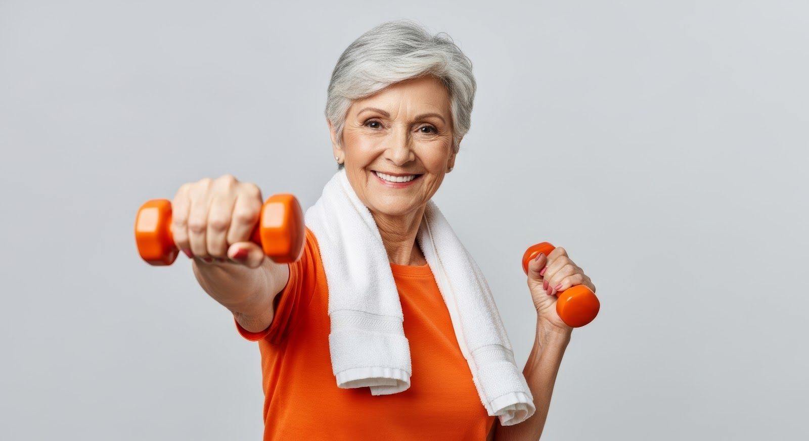 Smiling older woman in an orange top with a white towel around her neck lifting a pair of orange dumbbells