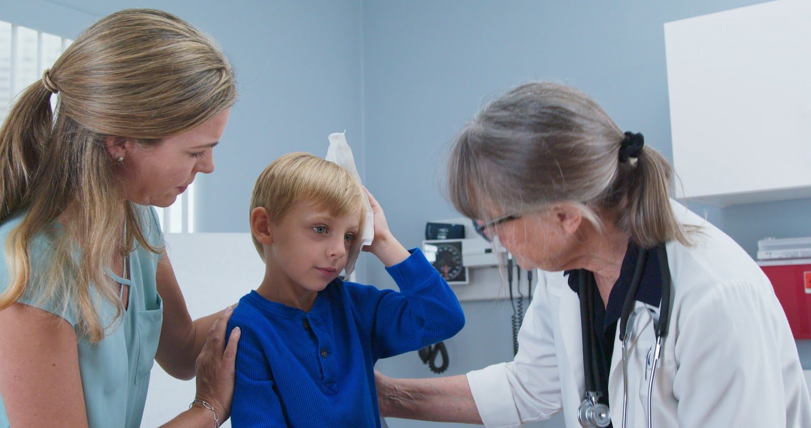Young boy in a blue shirt sitting in a clinic examination room holding an ice pack to his head, his mother beside him with a hand on his shoulder and a doctor in a white coat examining him