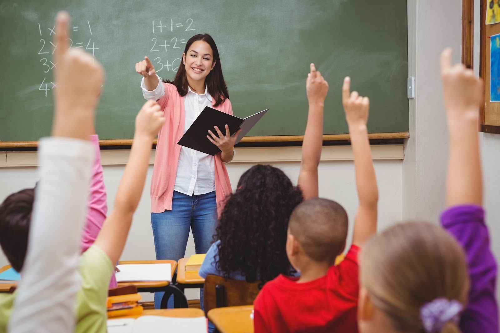 Teacher standing at a chalkboard holding an open book while young children in a classroom raise their hands