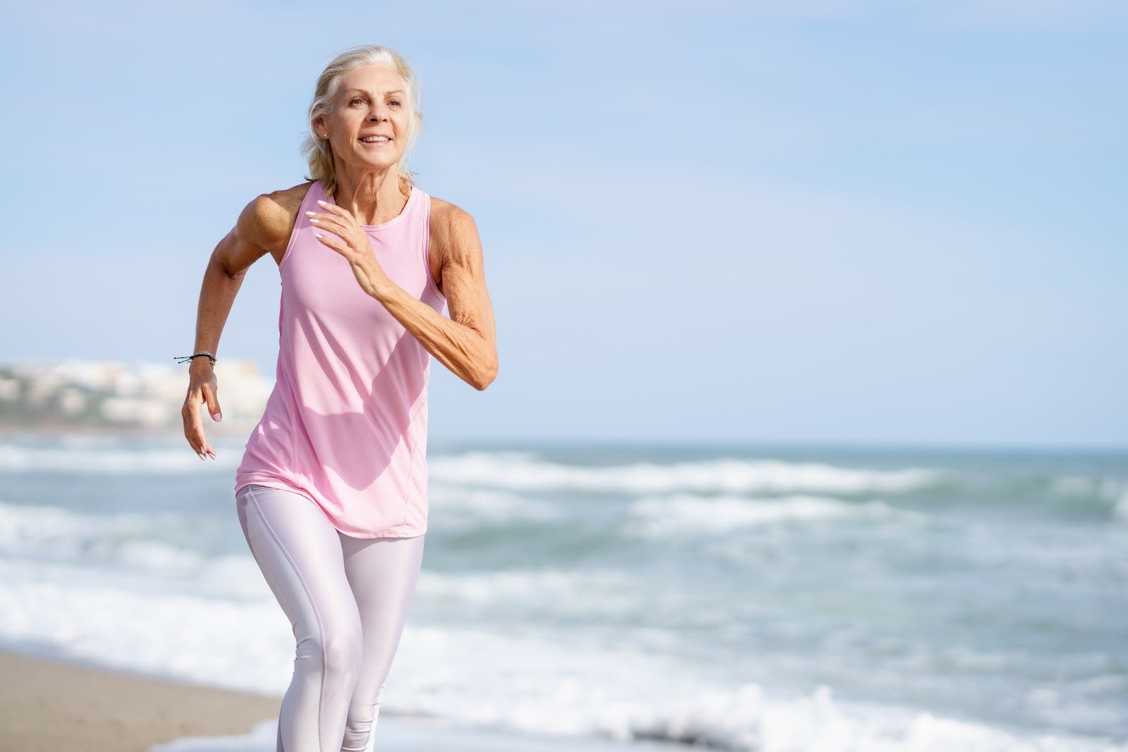 A mature woman in a pink top and white leggings running along the shoreline of a beach — the active, bone-and-muscle-strong later life that Nutrition 231 teaches chiropractors to support with nutrition by age, hormonal balance and osteoporosis care