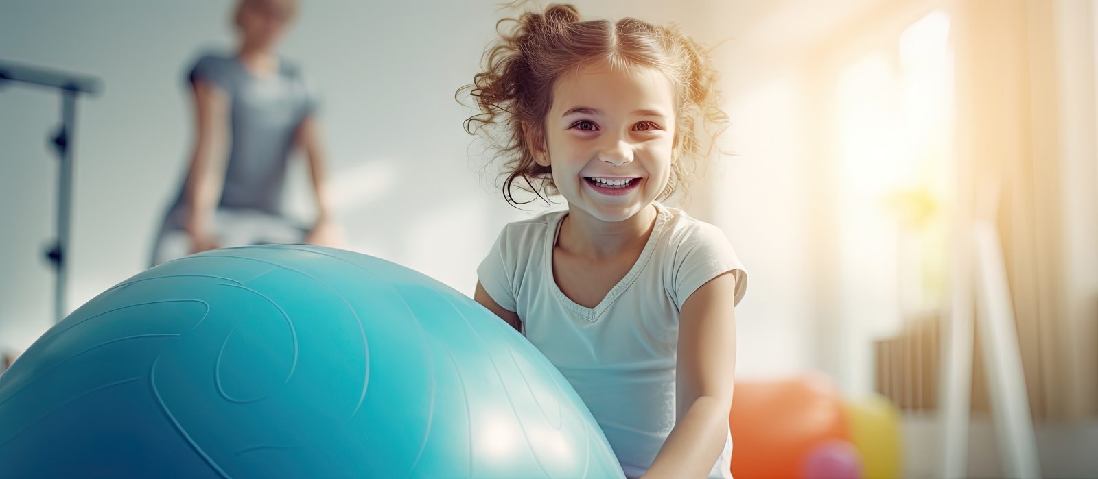 Smiling young girl holding a large teal therapy ball in a bright pediatric clinic, a clinician and second child behind her — pediatric adjusting and office procedures CE for chiropractors.