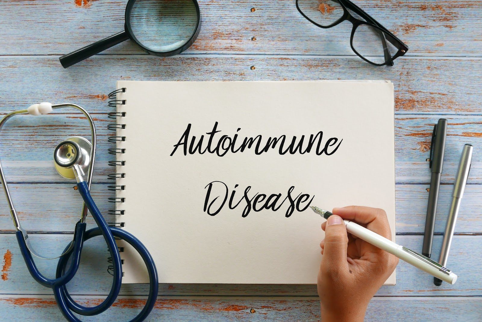 Notebook page with the words Autoimmune Disease handwritten beside a stethoscope, magnifying glass, glasses and a pen on a wooden desk