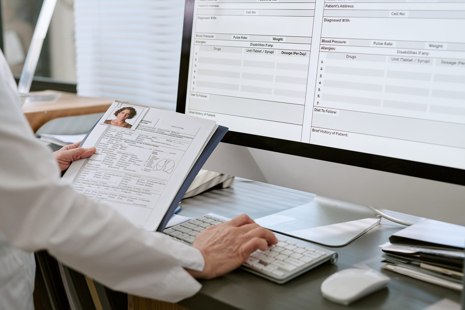 Clinician in a white coat holding a paper patient chart while entering data into an electronic health record on a desktop monitor — Documentation & Risk: Audit-Ready Workflows live chiropractic CE webinar with Dr. Michael Hall, CCEDseminars