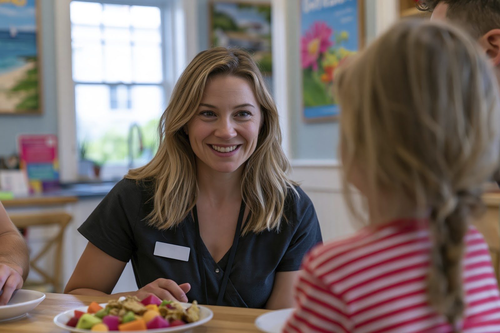 A smiling clinician in dark scrubs talking with a young girl with braided hair across a table holding a plate of fresh fruit in a bright clinic — the child-appropriate diet counseling that Nutrition 235 teaches chiropractors for pediatric nutrition