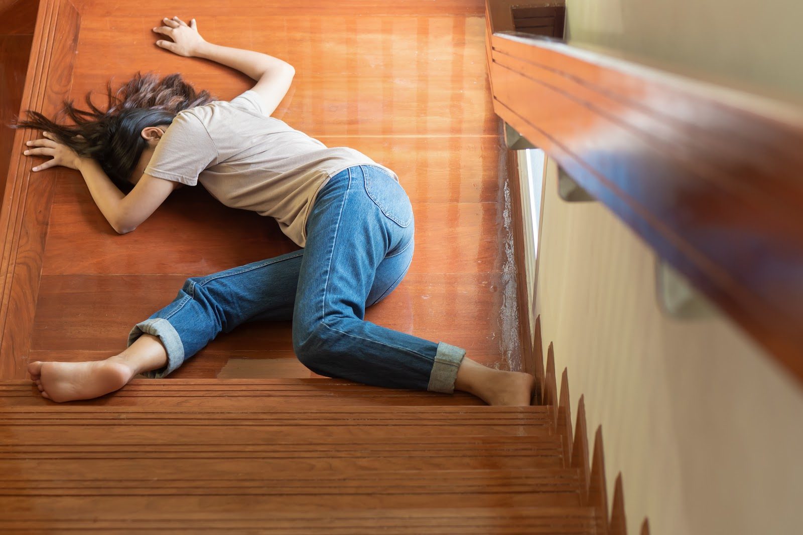 Person lying unconscious at the foot of a wooden staircase, viewed from above, after a collapse