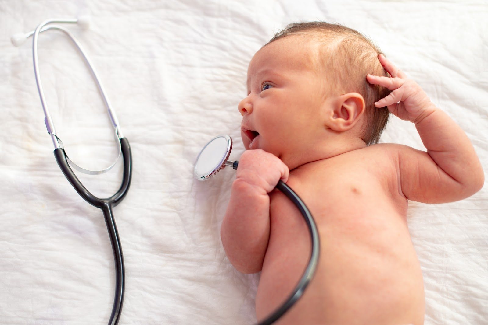 Newborn baby lying on white bedding, one hand raised near the head, with a stethoscope resting on the blanket alongside