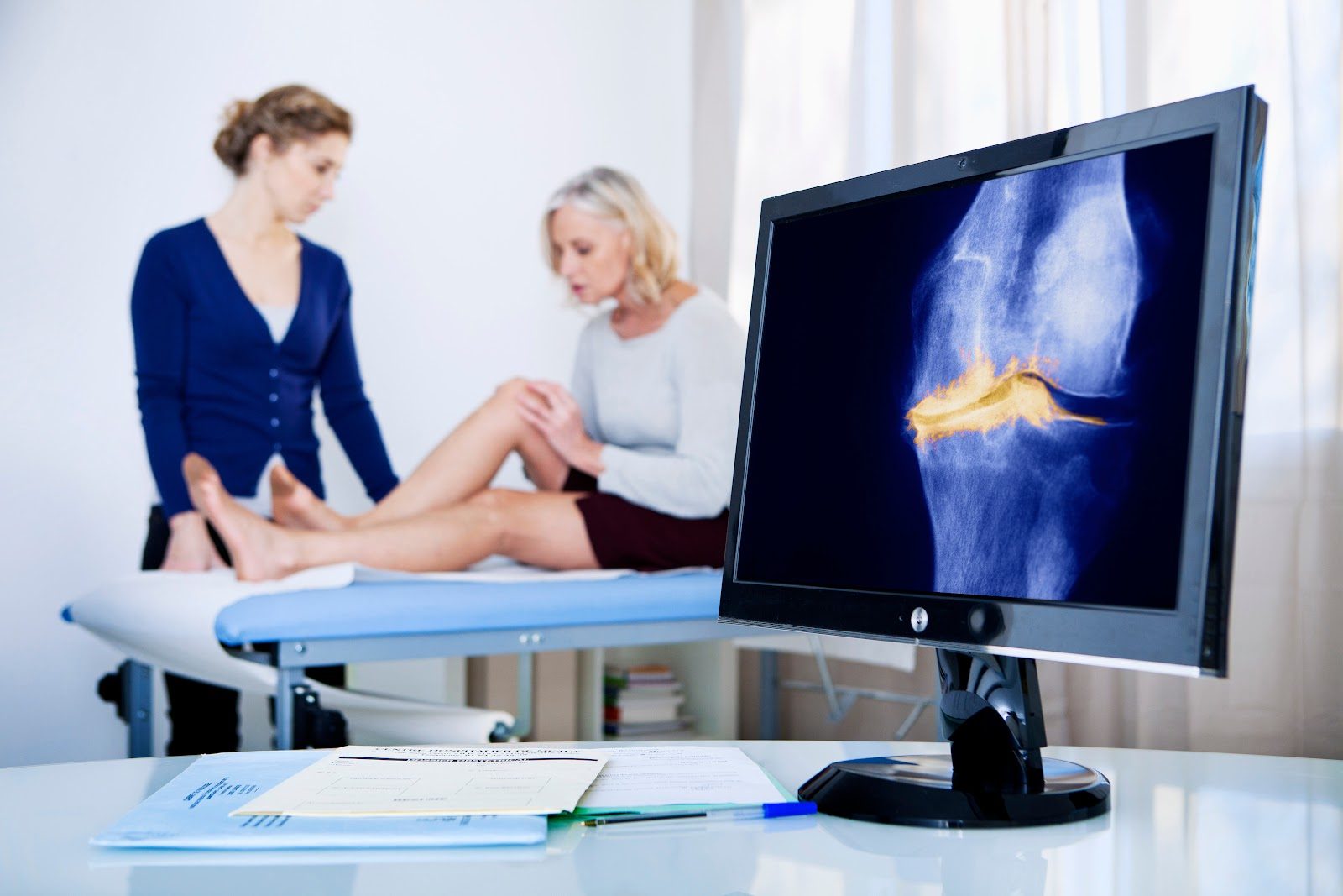 Clinician examining an older woman&rsquo;s knee on a treatment table while a monitor in the foreground displays a knee joint scan with the articular surfaces highlighted in yellow