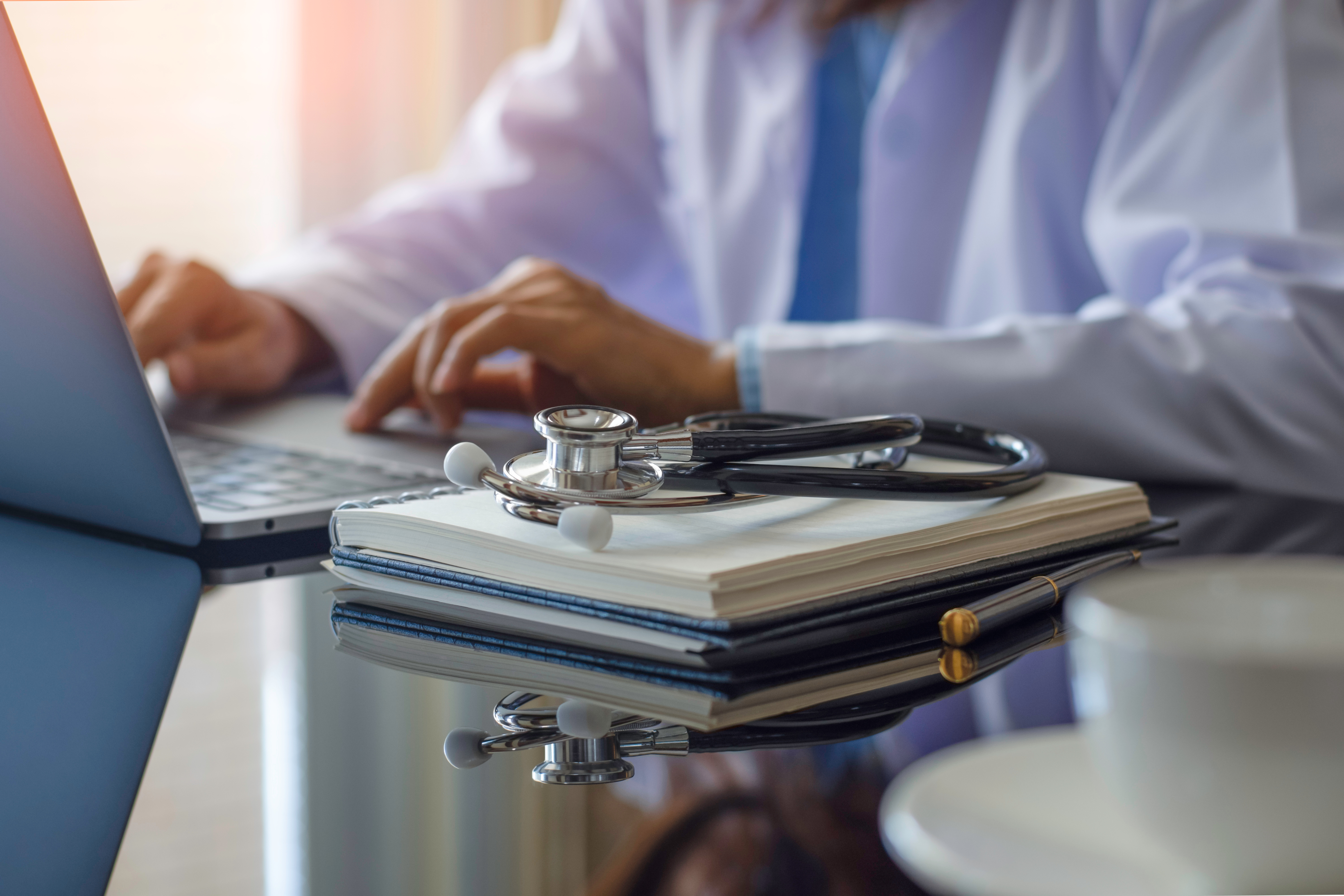 Chiropractor in white coat working at a laptop with a stethoscope resting on a clinical notebook — symbolizing CCEDseminars online chiropractic continuing education and renewal-ready course bundles