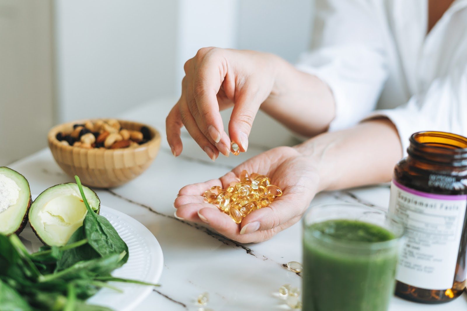 A clinician’s hands holding omega-3 and vitamin D capsules above a table of leafy greens, a green smoothie and a supplement bottle — the vitamin D testing, dosing and cofactor decisions that Nutrition 227 teaches chiropractors for bone and muscle health