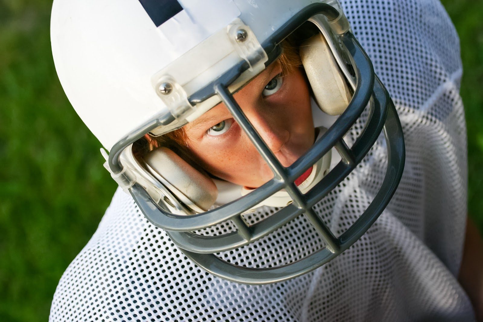 Close-up of a teenage American football player&rsquo;s face seen through the bars of a white helmet facemask, eyes fixed forward, shoulder pads and jersey visible