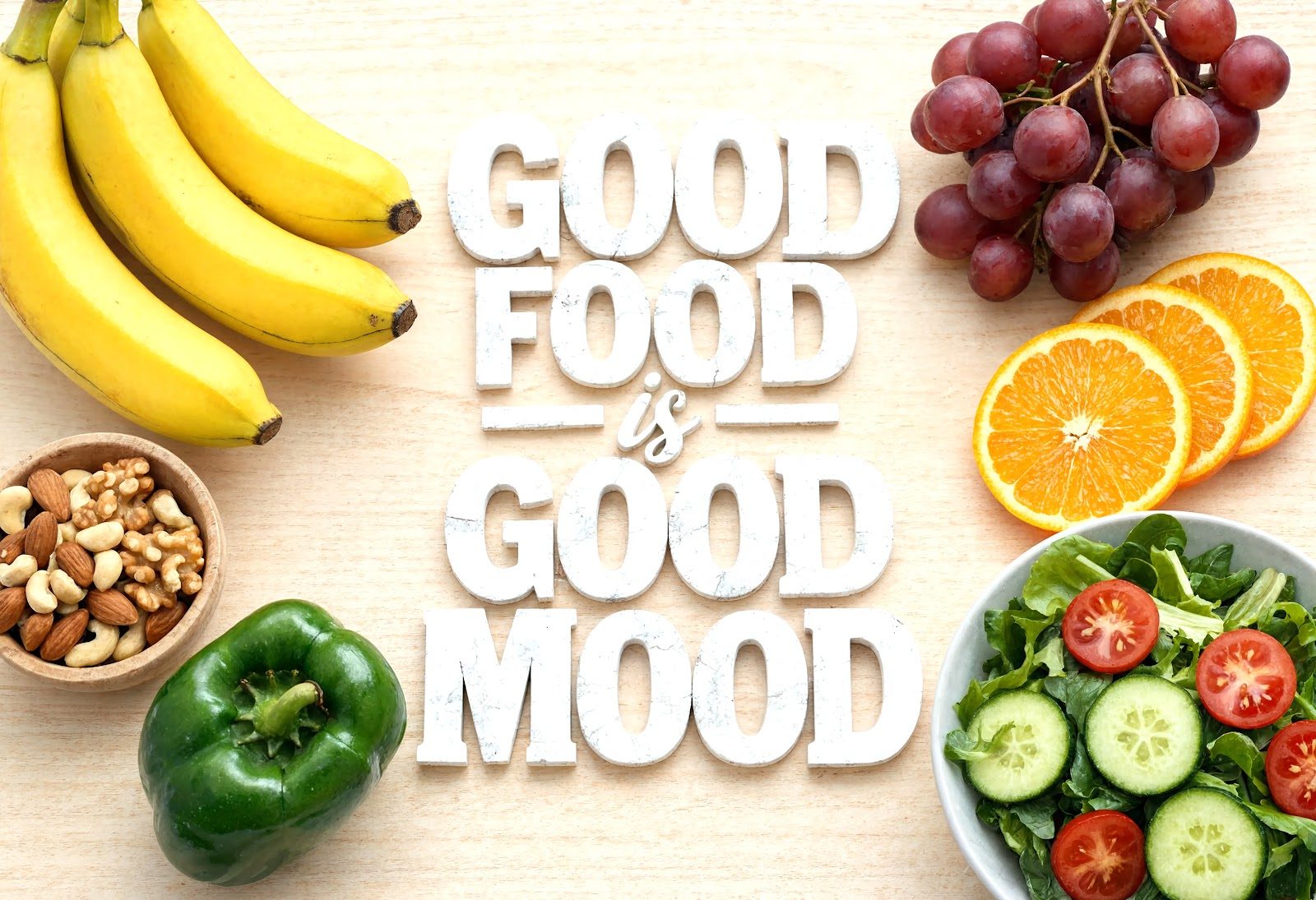 Flat-lay of bananas, grapes, oranges, mixed nuts, a green bell pepper and a cucumber-and-tomato salad around the words Good Food Good Mood — the diet-to-mood connection Nutrition 218 teaches chiropractors through neurotransmitter biochemistry