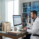 A chiropractor seated at a desk reviews patient documentation on an EHR screen in a modern clinical office, with printed charts and a spine model visible in the background.