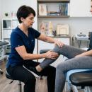 A chiropractor performs a hands-on orthopedic knee examination on a seated patient in a clinical setting, assessing joint function and range of motion.