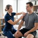 A sports medicine chiropractor performs a shoulder orthopedic examination on a seated male patient, assessing glenohumeral joint function in a clinical setting.