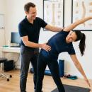 A chiropractor guides a patient through a coronal plane lateral bending movement in a clinical setting, demonstrating functional spinal mobility and neuroplasticity-based exercise.