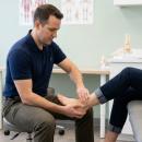 A chiropractor performs a hands-on foot and ankle orthopedic examination on a seated patient, assessing joint integrity and range of motion in a clinical environment.