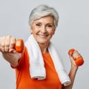 Smiling older woman in an orange top with a white towel around her neck lifting a pair of orange dumbbells