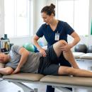 Clinician in navy scrubs examining the hip of a man lying on his side on a treatment table, knee flexed, in a bright rehab clinic with anatomy charts, resistance bands and exercise balls — the lumbopelvic-hip examination taught in Physical Therapy 205, a chiropractic CE course from CCEDseminars