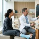 Clinician in a white coat pointing a pen at bilateral ankle radiographs on a wall-mounted monitor while a seated patient in a blue short-leg cast looks on, an ankle model on the desk — the ankle and lower leg examination, the subject of Physical Examination 206: Knee and Lower Leg, a two-hour chiropractic CE course from CCEDseminars