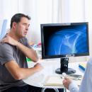Patient in a gray T-shirt holding his painful shoulder and describing the complaint while a clinician in a white coat reviews a shoulder radiograph on a monitor between them — the shoulder complex examination, the subject of Physical Examination 209: Shoulder Complex, a two-hour chiropractic CE course from CCEDseminars