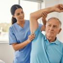 Clinician in blue scrubs guiding an older patient's arm overhead, one hand supporting the elbow and the other stabilizing the shoulder, in a bright exam room — the upper extremity examination, the subject of Physical Examination 210: Upper Extremity, a two-hour chiropractic CE course from CCEDseminars